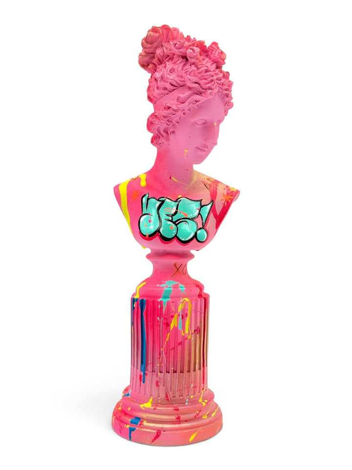 A vibrant pink classical female bust sculpture on a fluted pedestal base, decorated with graffiti-style bubble lettering and paint splatters in yellow, blue, and green, set against a plain white background.